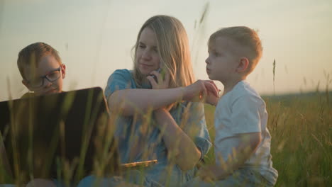 a mother in a blue gown multitasks in a grassy field, using a laptop and phone, with her two young sons beside her, one in yellow engrossed in the laptop,the other in white,playfully touching her hair