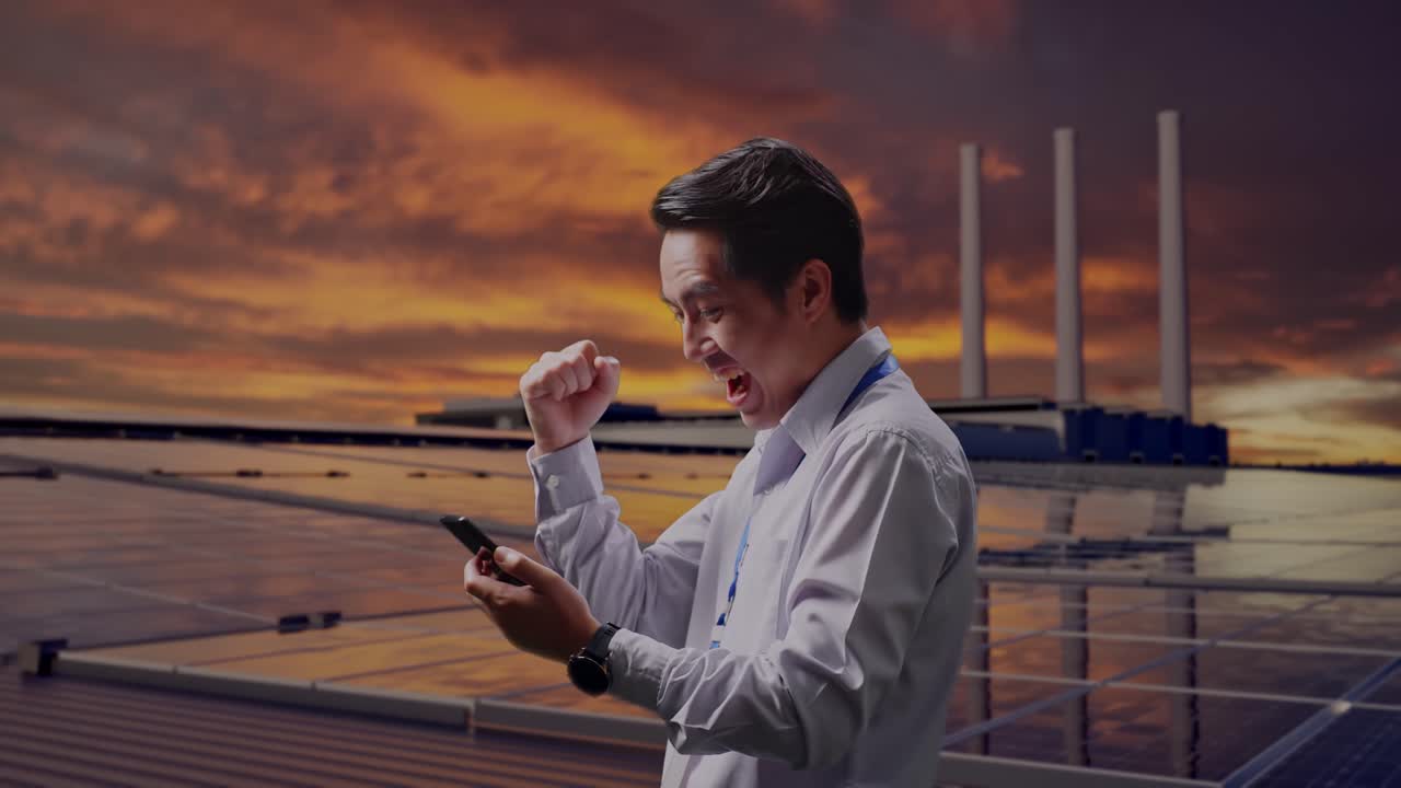 Vista laterale di un lavoratore professionista asiatico con il suo smartphone, ispeziona pannelli solari su un tetto al tramonto, esulta a pugno alzato dopo aver controllato lo smartphone.