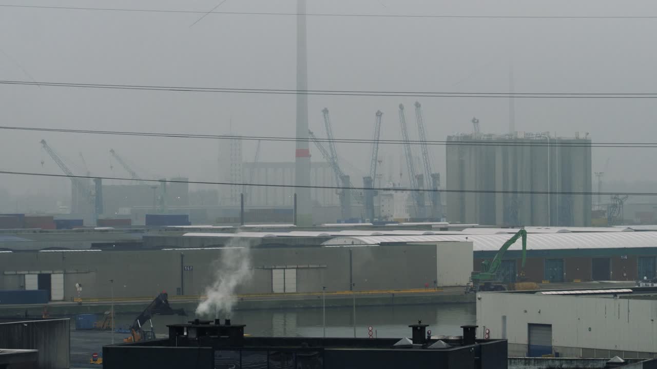 A misty harbor with industrial cranes, silos, and waterfront buildings, with smoke rising from a chimney under an overcast sky, creating a calm industrial scene perfect for trade and logistics themes