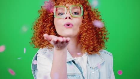 joven pelirroja juguetona mujer rizada soplando confeti colorido a la cámara, sonriendo sobre el fondo clave de croma verde