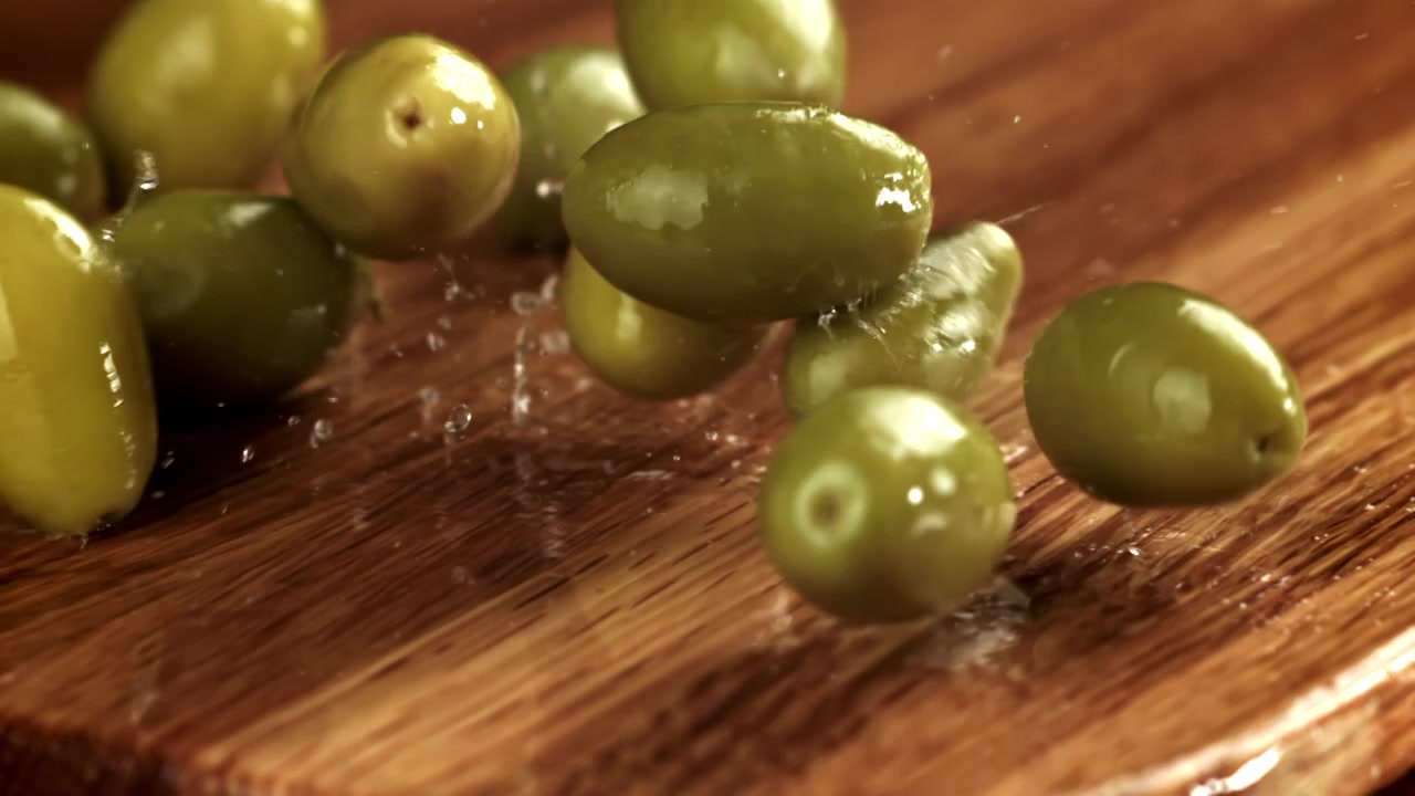 Olive verdi che cadono e schizzano acqua su un tagliere di legno al rallentatore