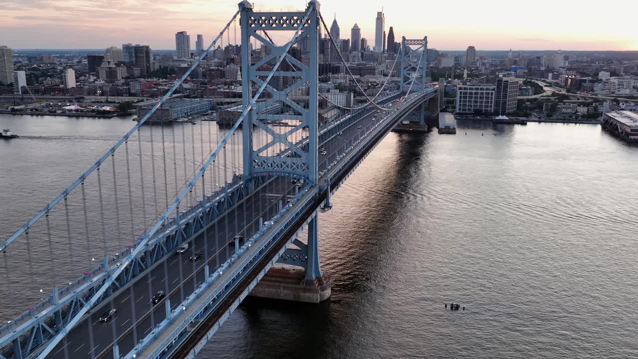 Aerial flight over Delaware River passing benjamin Franklin bridge in Philadelphia. Skyline with skyscrapers in distance. Wide shot. Peaceful sunrise in morning. Cars on bridge