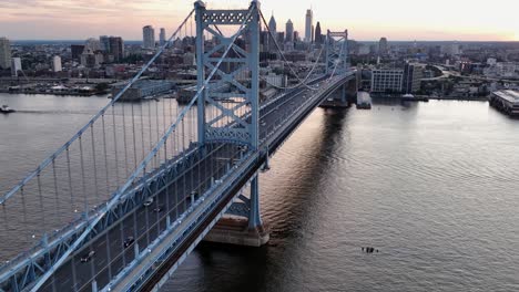 aerial flight over delaware river passing benjamin franklin bridge in philadelphia. skyline with skyscrapers in distance. wide shot. peaceful sunrise in morning. cars on bridge