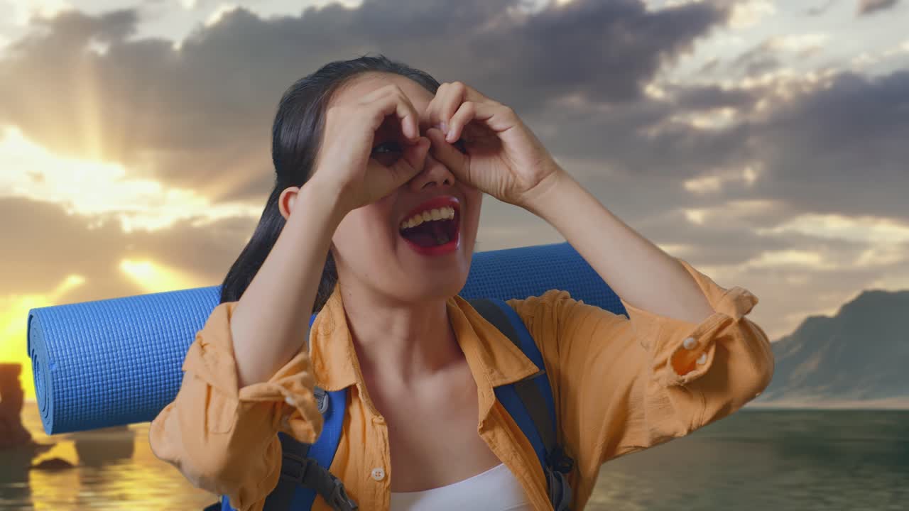 Primo piano di escursionista asiatica con zaino da alpinismo che sorride e fa il gesto del binocolo, poi si guarda intorno mentre è in piedi sulla spiaggia durante il tramonto