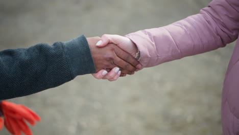 Close-up of extended hand in pink winter jacket reaching towards another hand in dark sleeve holding orange gloves, symbolizing approach, greeting, handshake, agreement, cooperation in outdoor setting