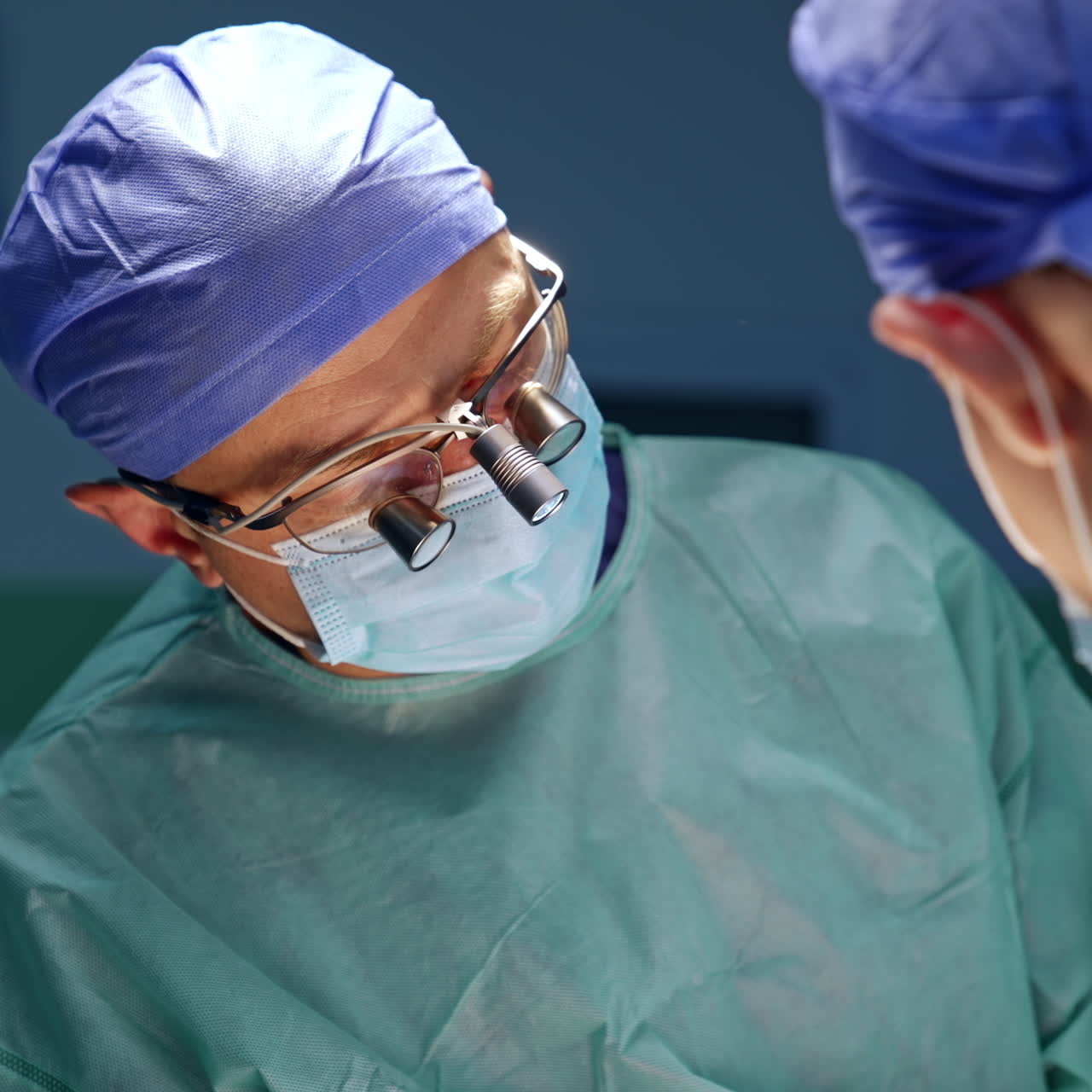 Chirurgo con loupes chirurgiche sta in piedi con la testa inclinata a destra, concentrato sul campo operatorio. Operazione in corso.