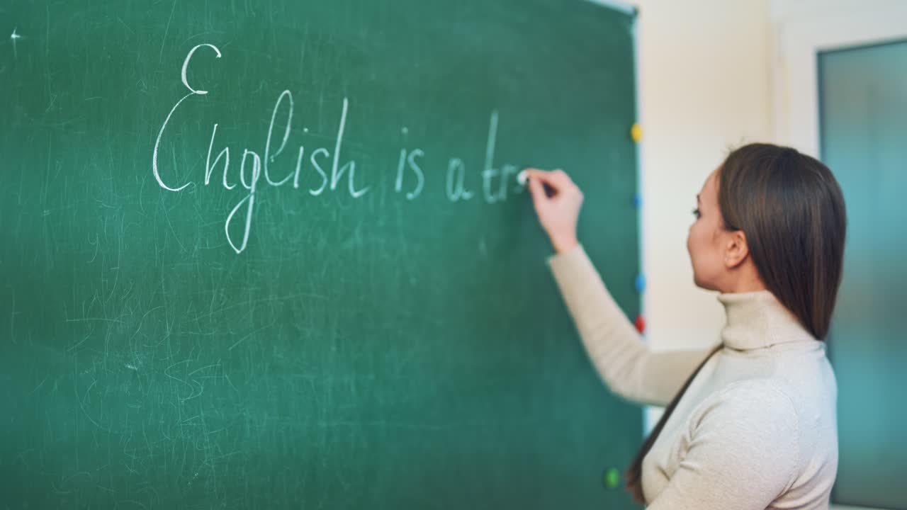 Young pedagogue writes on a blackboard with a chalk in the educational center. Attractive teacher is writing English words on a board in the classroom.
