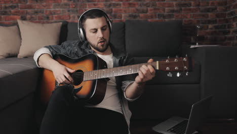 joven músico tocando la guitarra y usando una laptop en casa