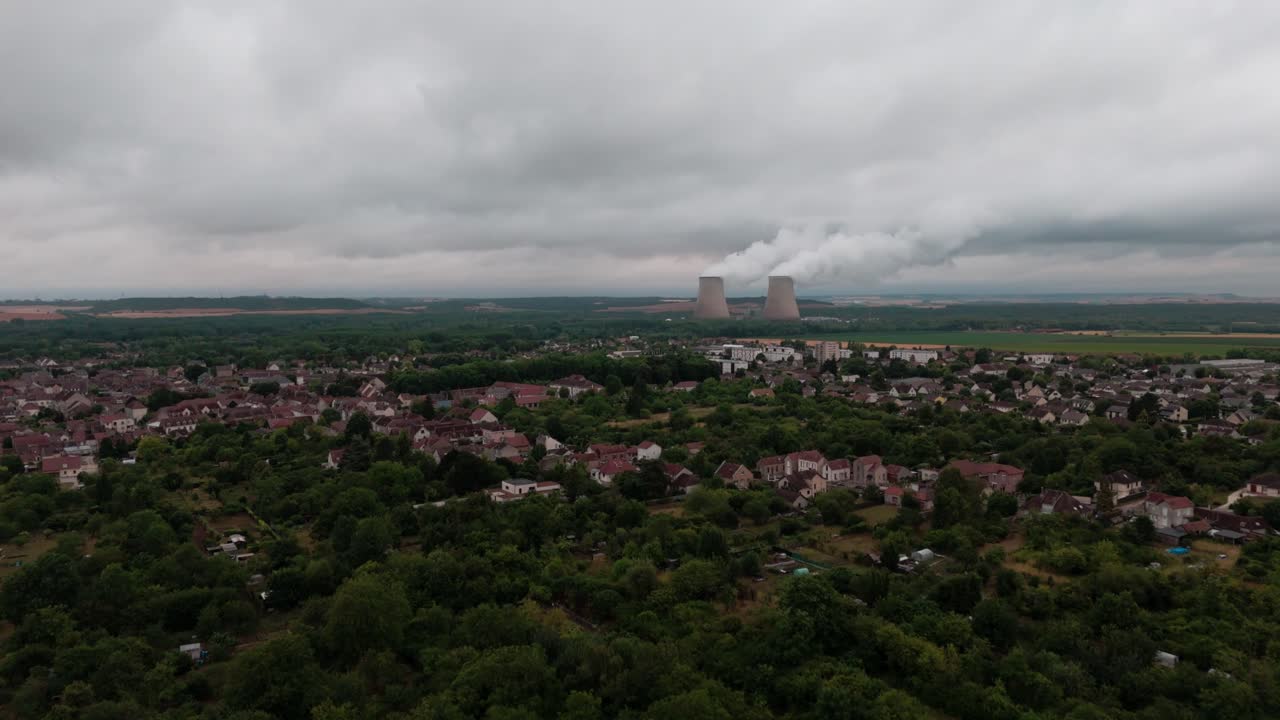 French village with nuclear power plant on horizon, towers emitting steam, overcast sky, France. Aerial forward, copy space