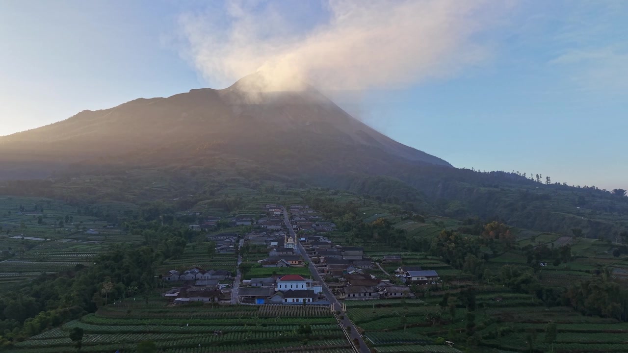 Ripresa aerea di un insediamento rurale indonesiano sotto il Monte Merapi. Il paesaggio montano vulcanico e i terreni agricoli offrono uno scenario visivo mozzafiato.