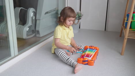 Mixed-Ethnicity-Toddler-of-Asian-Descent-Sitting-on-Floor-Playing-with-Toy-Xylophone-at-Home-Balcony