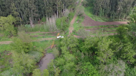 aerial view of a white pickup truck crossing a stream in a rural area near a forest, with a small wooden bridge visible in the background