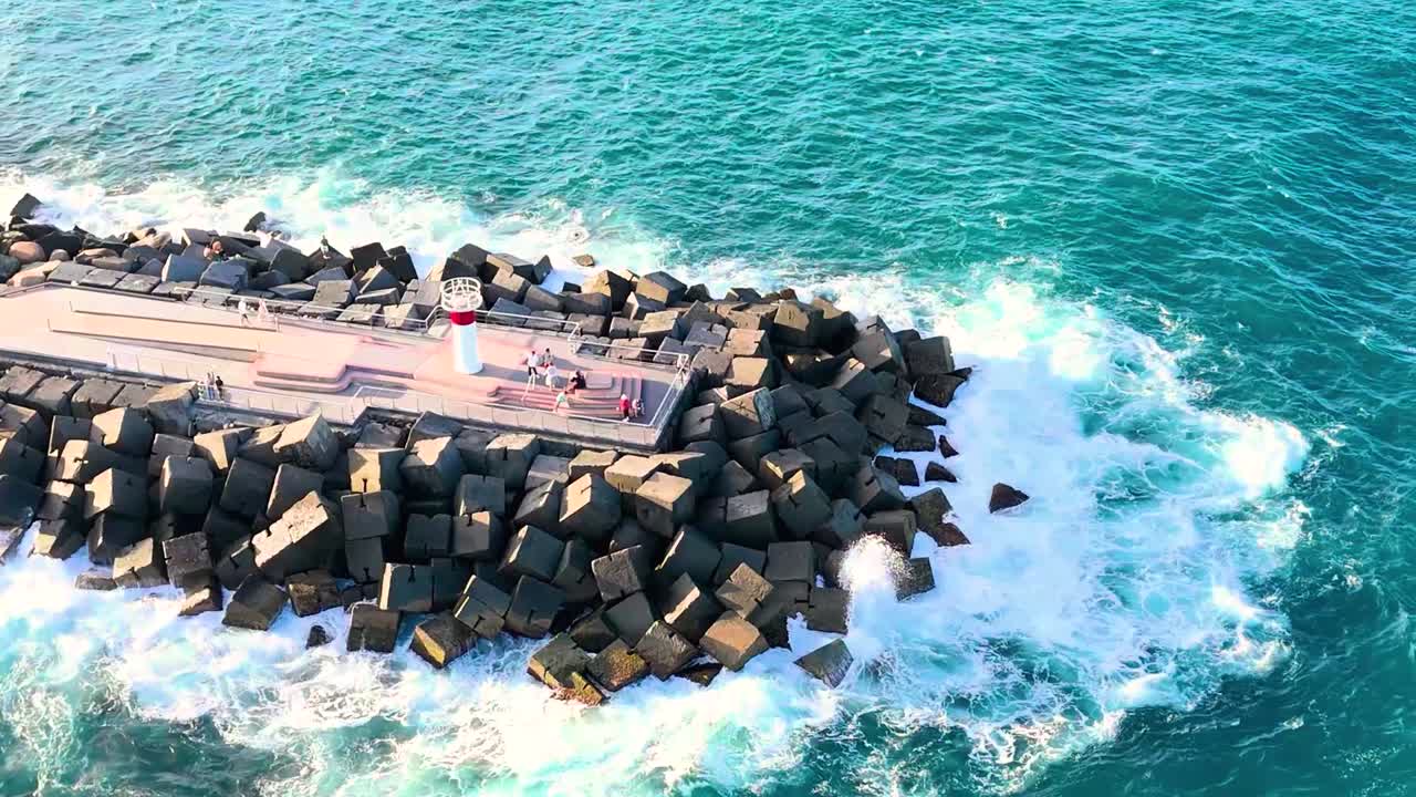 Aerial view of ocean waves hitting a rock jetty with a walkway and lighthouse.