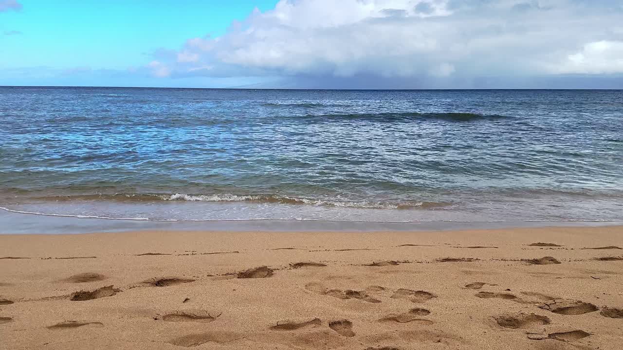Vlny narážející na písečnou pláž s modrou oblohou a lehkými mraky, pořízeno na Maui, Havaj.