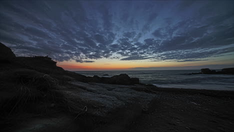 Timelapse-De-ángulo-Bajo-En-La-Playa-Con-Puesta-De-Sol-Bajando-En-El-Horizonte-Sobre-El-Mar-Con-Nubes-Pasando-Volando