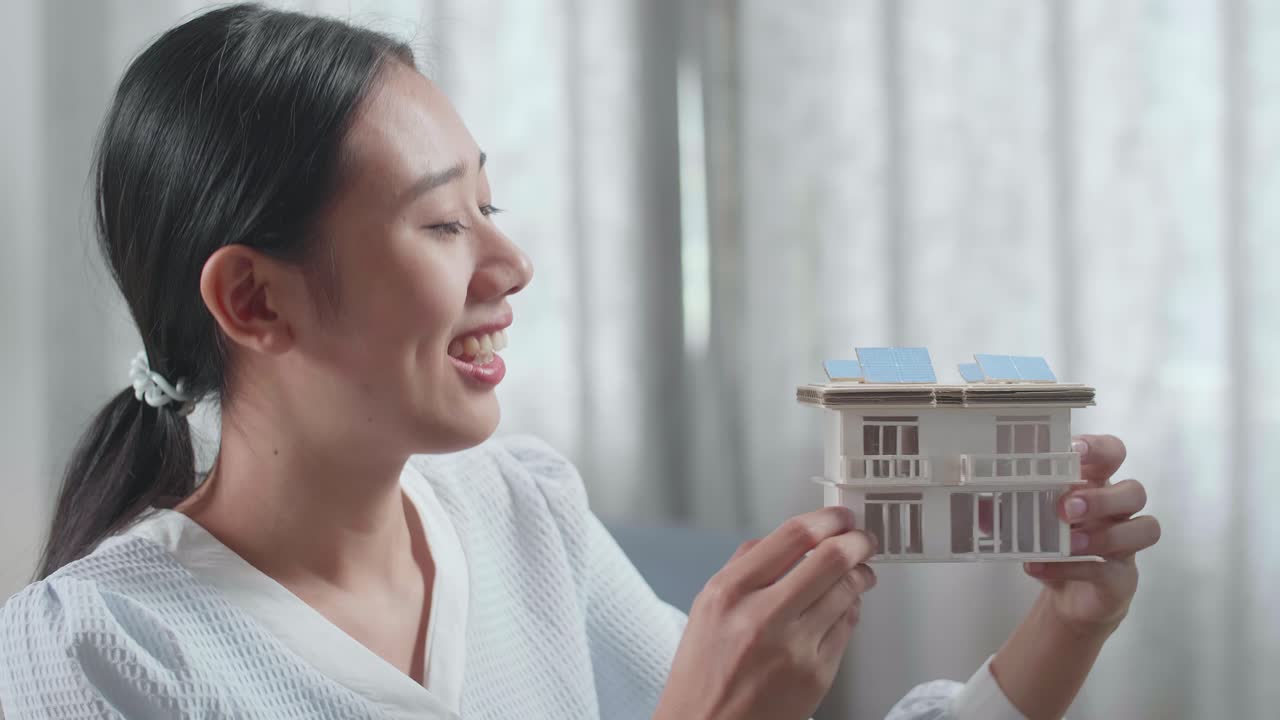 아시아 여성 엔지니어가 집에 있는 데스크에 있는 사진과 비교하기 전에 태양광 패널이 있는 작은 집 모델을 들고 있는 모습입니다.