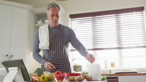 Hombre-Caucásico-Feliz-Usando-Delantal,-Parado-En-La-Cocina,-Cocinando-La-Cena-Y-Usando-Una-Tableta