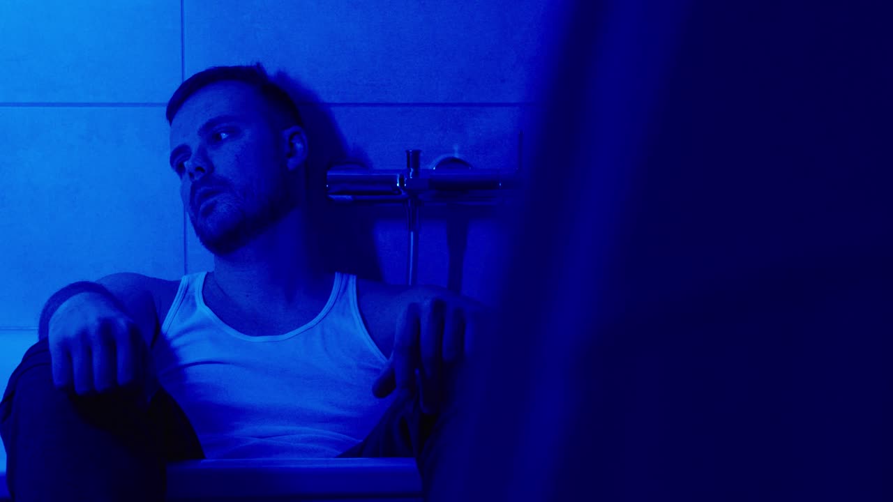 Young man sitting in a bathtub at night under blue light, looking emotionally drained while using his smartphone. Concept for loneliness, depression, digital addiction, and mental health