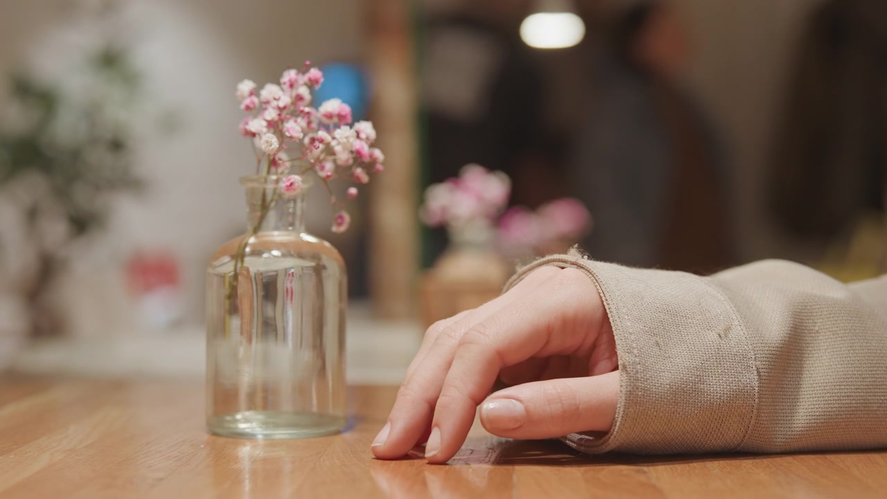 Gros plan sur une main posée sur une table en bois près d'un vase en verre transparent avec des fleurs roses. L'arrière-plan montre un espace intérieur flou avec un éclairage doux et des arrangements floraux supplémentaires. La main reste.