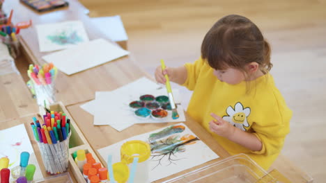 Linda-Niña-Sentada-Junto-Al-Escritorio-Usando-Pincel-Para-Dibujar-Y-Pintar-En-El-Papel