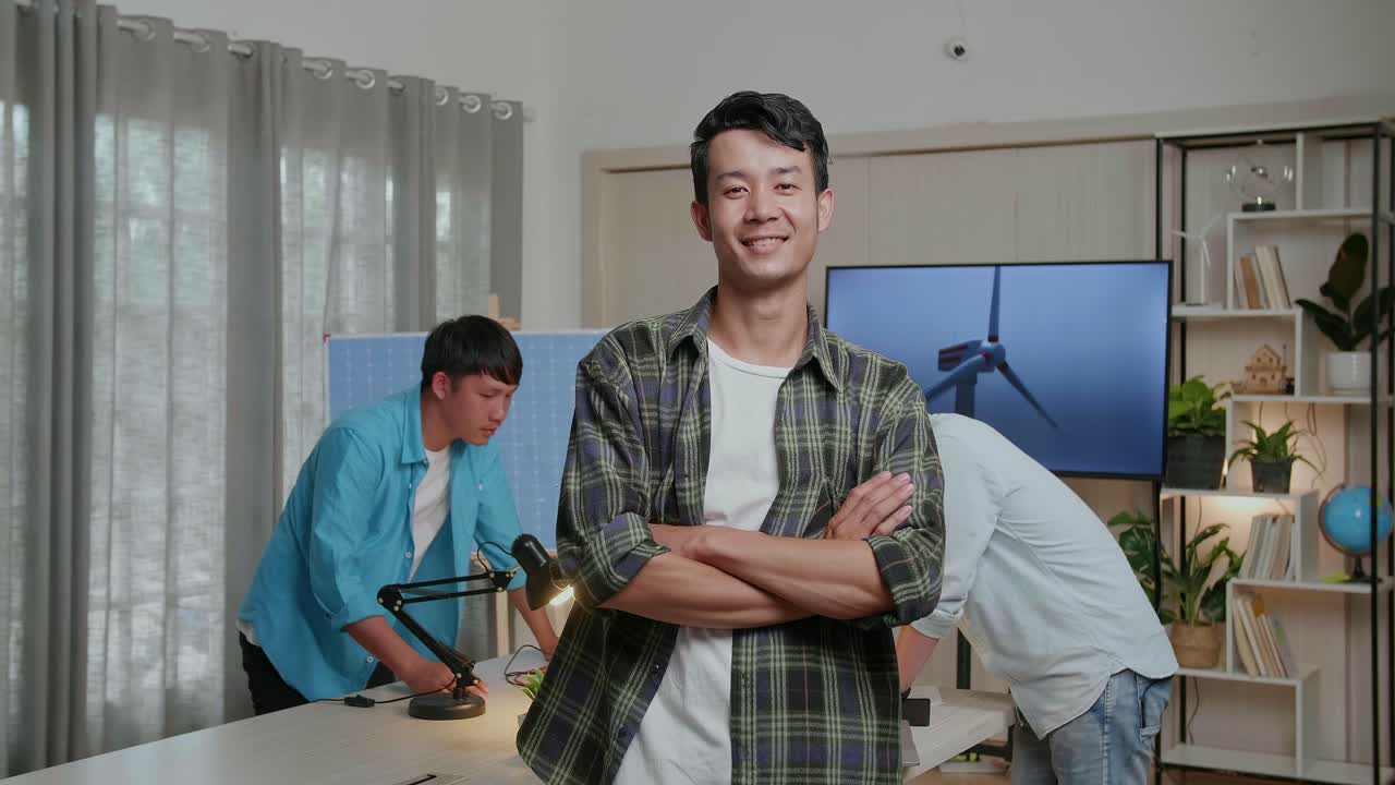 동료들이 사무실에서 풍력 터빈에 대해 논의하는 동안 미소 짓는 아시아 남자 엔지니어가 카메라에 팔을 고 있습니다.
