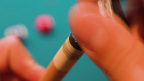 person chalking tip of pool or snooker cue with blue chalk on wooden stick closeup using grinding and brushing motion with pool balls and blue felt in background, no faces, billiards