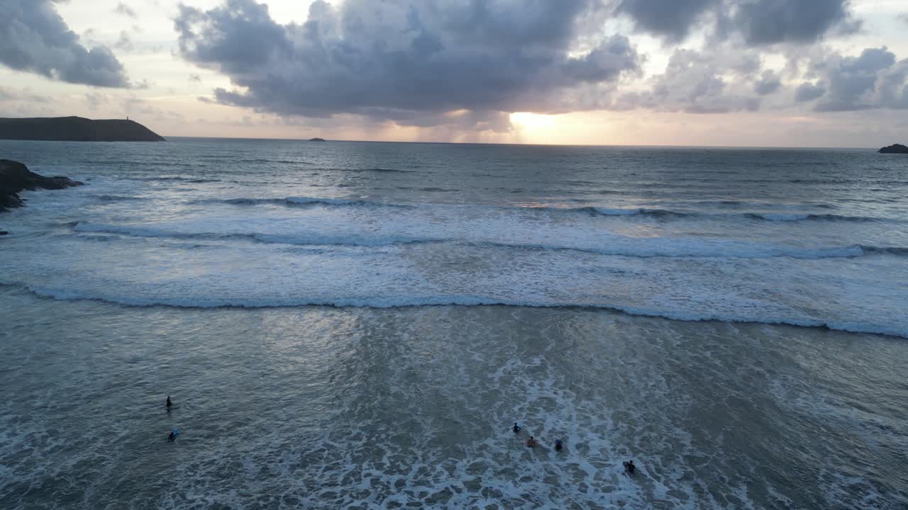 Un video aereo realizzato con un drone sopra la spiaggia di Polzeath cattura le serene onde dell'oceano al tramonto con nuvole suggestive. La scena tranquilla mette in risalto la bellezza del paesaggio costiero e il cielo vivace.