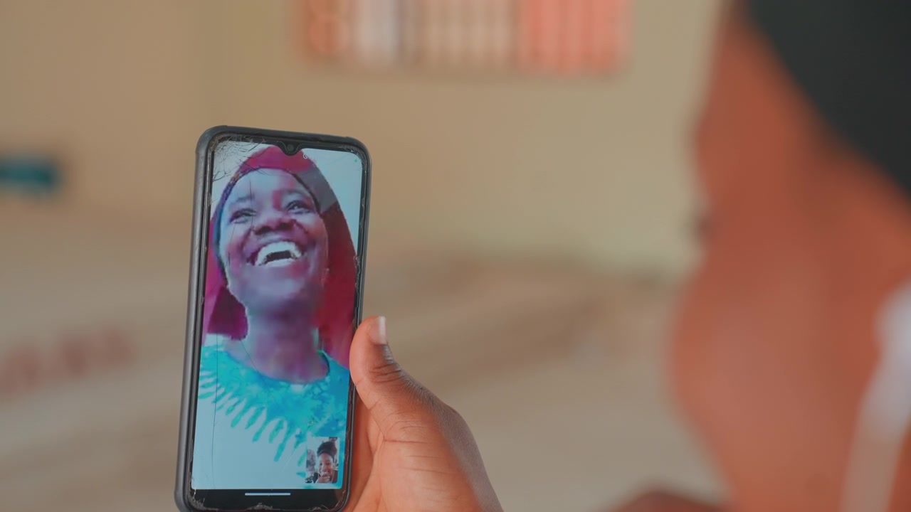 Bambino videochiama la madre sorridente al telefono, madre nera sorride sullo schermo mentre il bambino tiene il dispositivo, luce interna calda e intimo legame a distanza, ritratto spontaneo di connessione familiare
