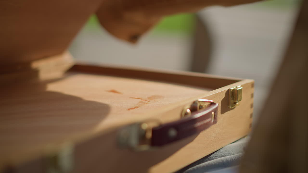 mani che chiudono una scatola di vernici di legno con chiusure in ottone e manico in pelle, la luce del sole proietta ombre sulla venatura, un leggero clic assicura il kit, rituale di fine sessione, atmosfera calma e slow living, attenzione all'hardware e alla texture