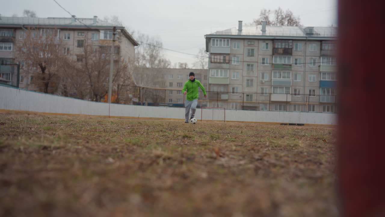 Beyaz bir adam, boş bir şehir sahasında uzun bir şut çekmek için uzaktan yaklaşıyor; kahverengi çimlere ve uzaktaki paslı kaleye karşı neon yeşili kapüşonlu sweatshirt'ü görülebiliyor, düşük kamera açısı hareketi vurguluyor
