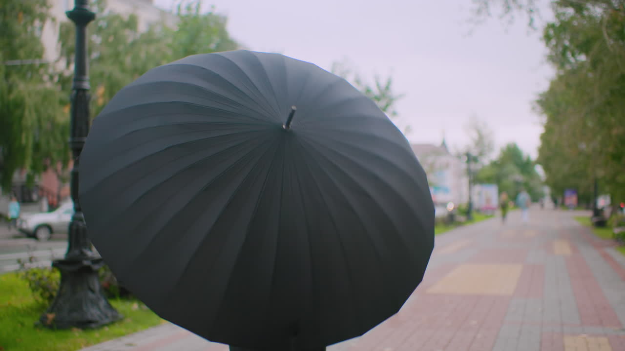 흐린 날씨에 도시 거리에서 검은 우산을 돌리며 서 있는 여성이 자동차, 건물, 나무의 흐릿한 배경 뒤에 숨어 있는 모습으로, 도시 라이프스타일의 야외 분위기를 나타내요.