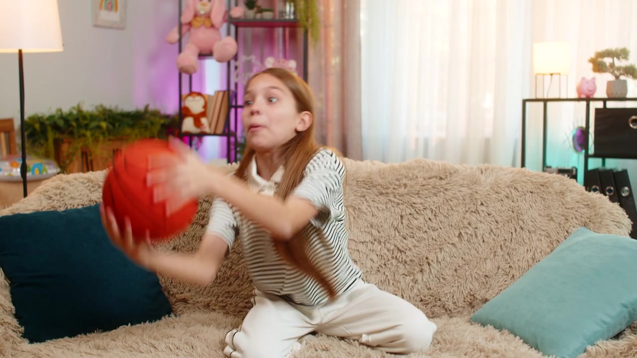 Child girl kid playing alone on sofa at home tossing basketball ball in air enjoying solo indoor fun