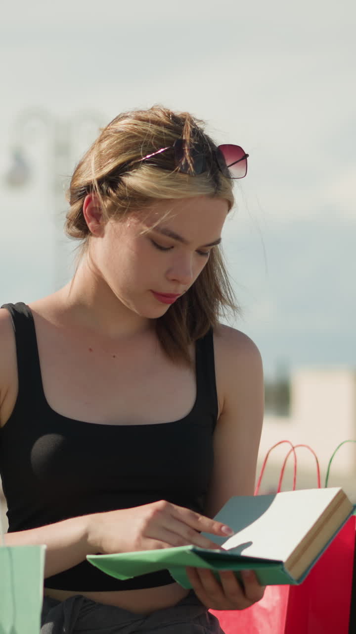 una señora sentada en un banco al aire libre con bolsas de compras a ambos lados recupera un libro de color menta de la bolsa verde menta, lo abre y hojea sus páginas, mientras que el fondo permanece ligeramente borroso