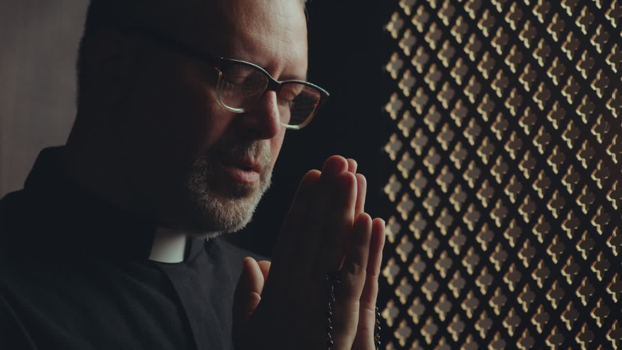 Priest Praying with Rosaries in Clasped Hands inside of Confessional