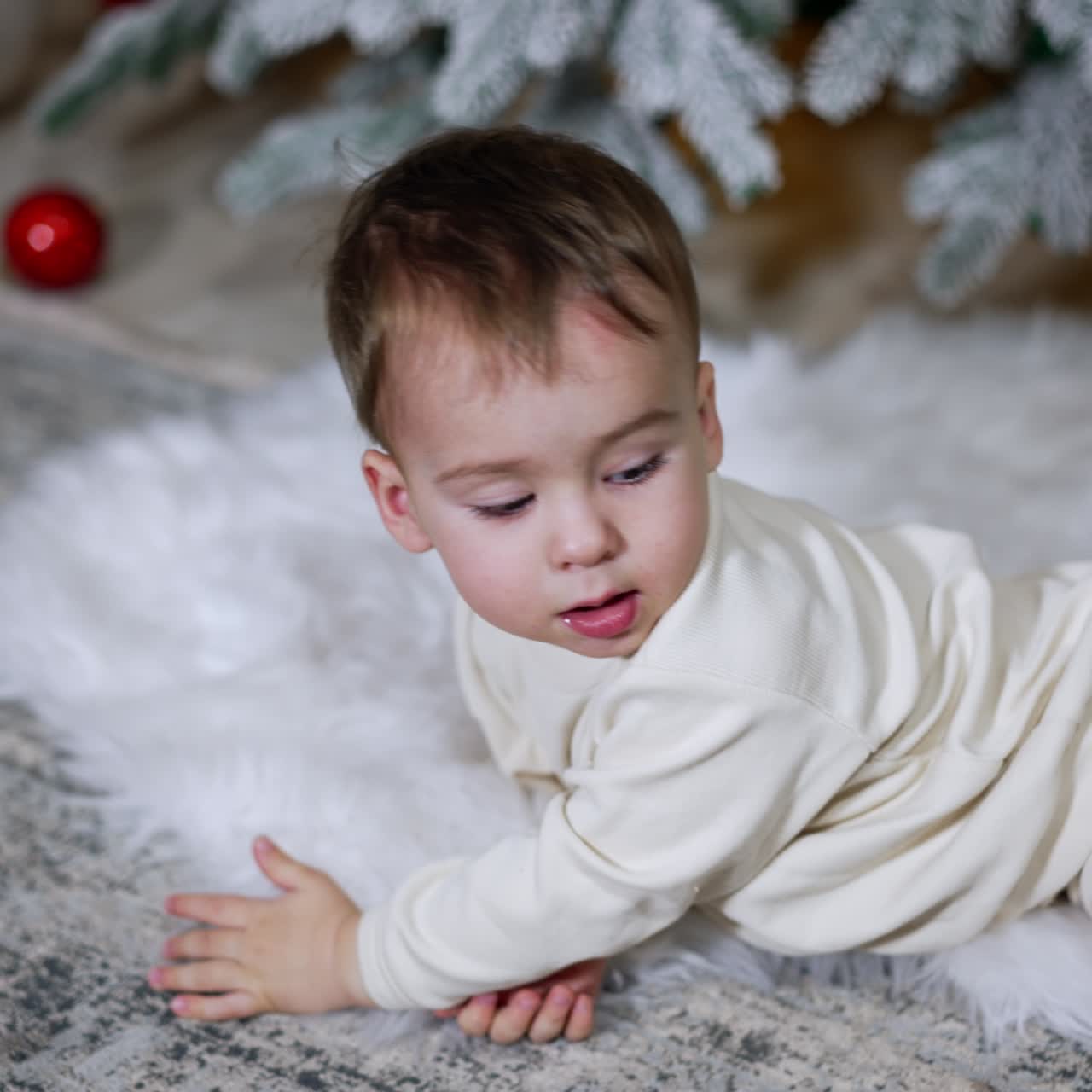 Angelic kid in white clothes lies on a fluffy soft plaid under the Christmas tree. Happy baby plays with a toy