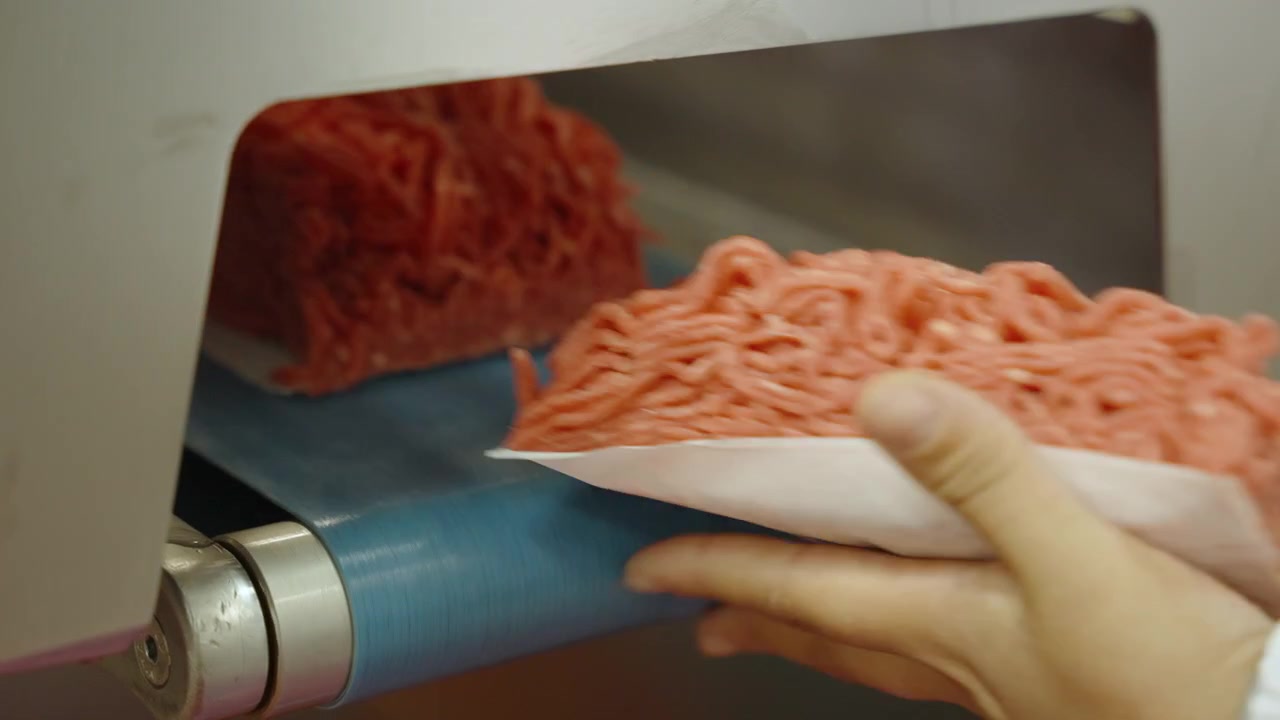 Des blocs de bœuf haché formés sortent d'une unité de formage sur un tapis bleu, tandis qu'une main gantée recueille chaque portion dans une usine de viande sud-africaine