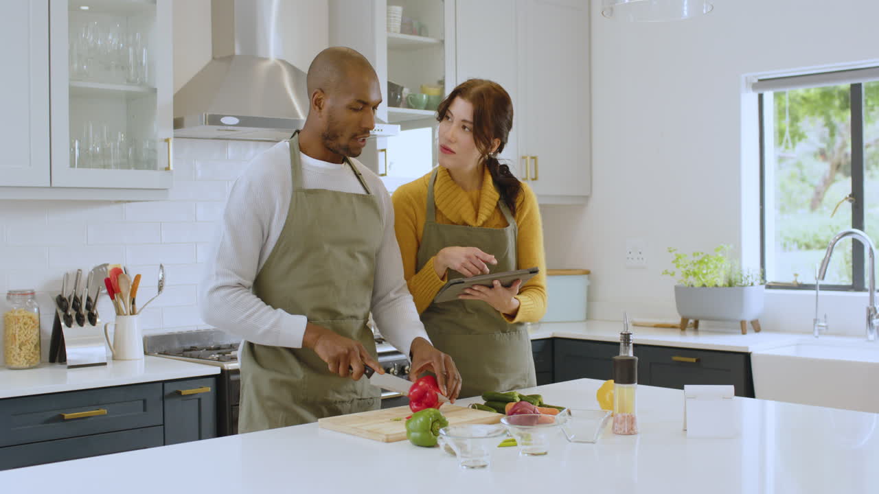 Multiracial couple cooking together in modern kitchen, using tablet for recipe guidance, at home