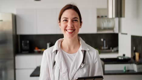 Portrait-of-a-happy-brunette-girl-in-a-white-doctors-coat-with-a-stethoscope-on-her-shoulders-and-with-a-tablet-in-her-hands-near-a-modern-kitchen-in-an-apartment-for-a-home-examination