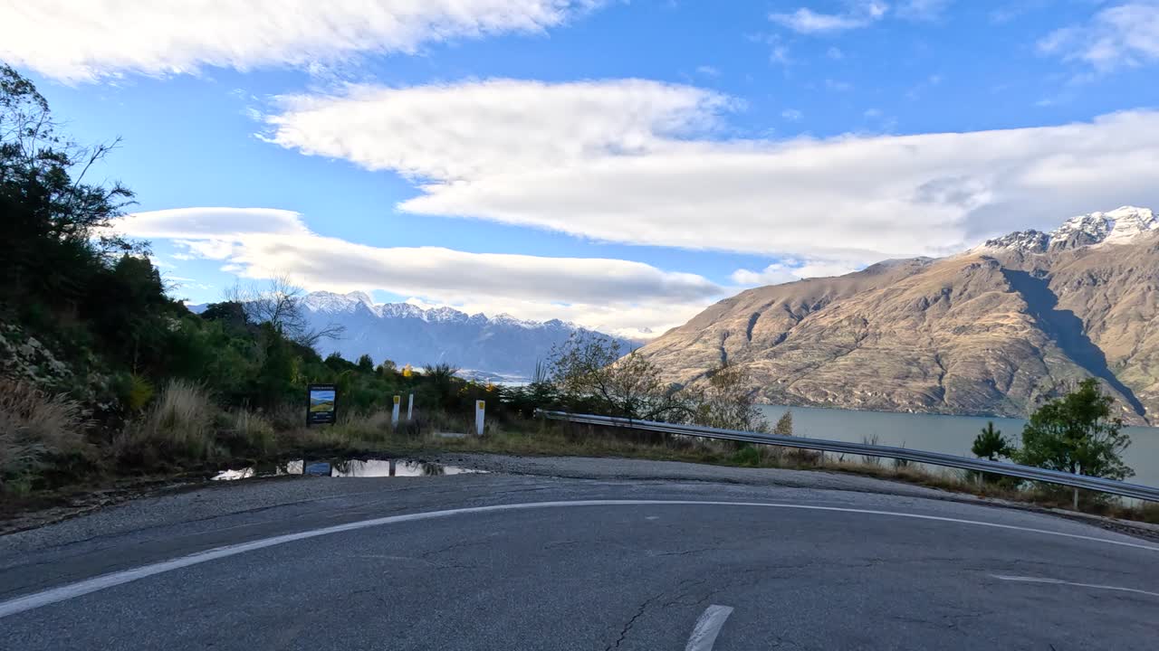 Vehicle drives winding lakeside road with mountain views, daylight, smooth camera movement, vibrant landscape