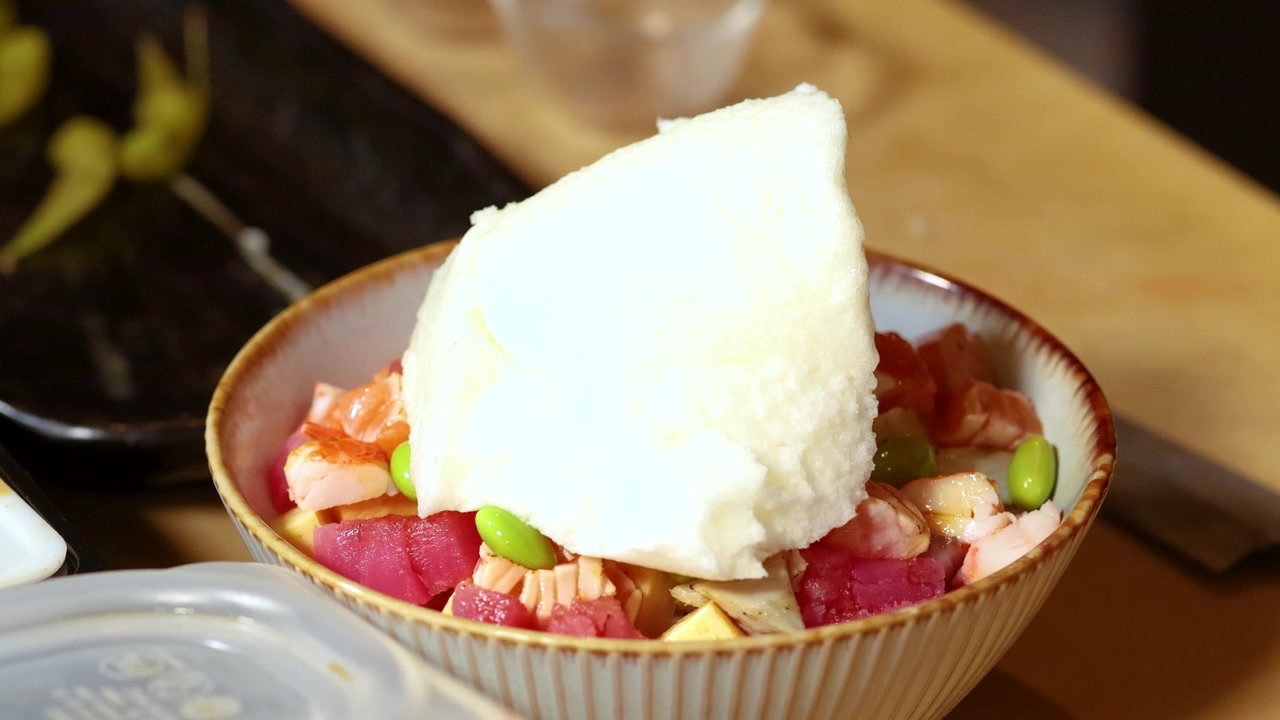 Close-up sendok yang menempatkan meringue putih ke dalam mangkuk poke ikan mentah berwarna-warni.