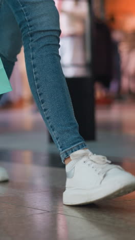 vista de la pierna de una persona con vaqueros y zapatillas de deporte de lona blanca sosteniendo bolsas de compras rosadas y menta mientras camina a través de un centro comercial más allá de una tienda de ropa, con fondo borroso de pantallas minoristas