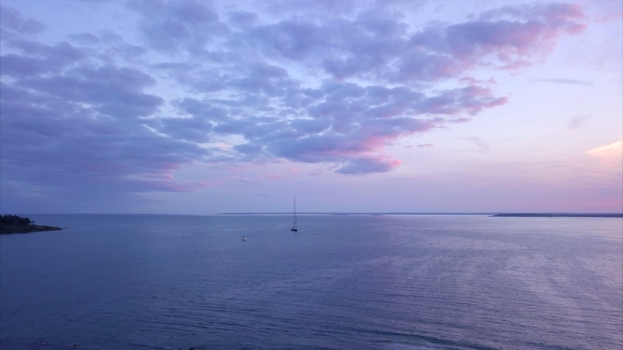 메인 해안선에서 극적인 분홍색과 보라색 일몰 동안 한 척의 범선으로 잔잔한 항구 위로 날아가는 공중 샷