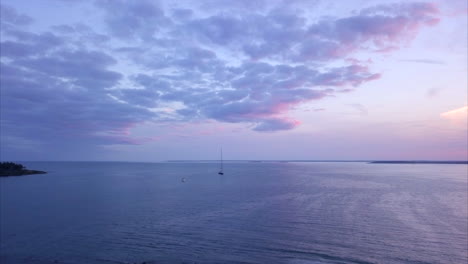 aerial shot flying out over a calm harbor with a single sailboat during a dramatic pink and purple sunset off the maine coastline