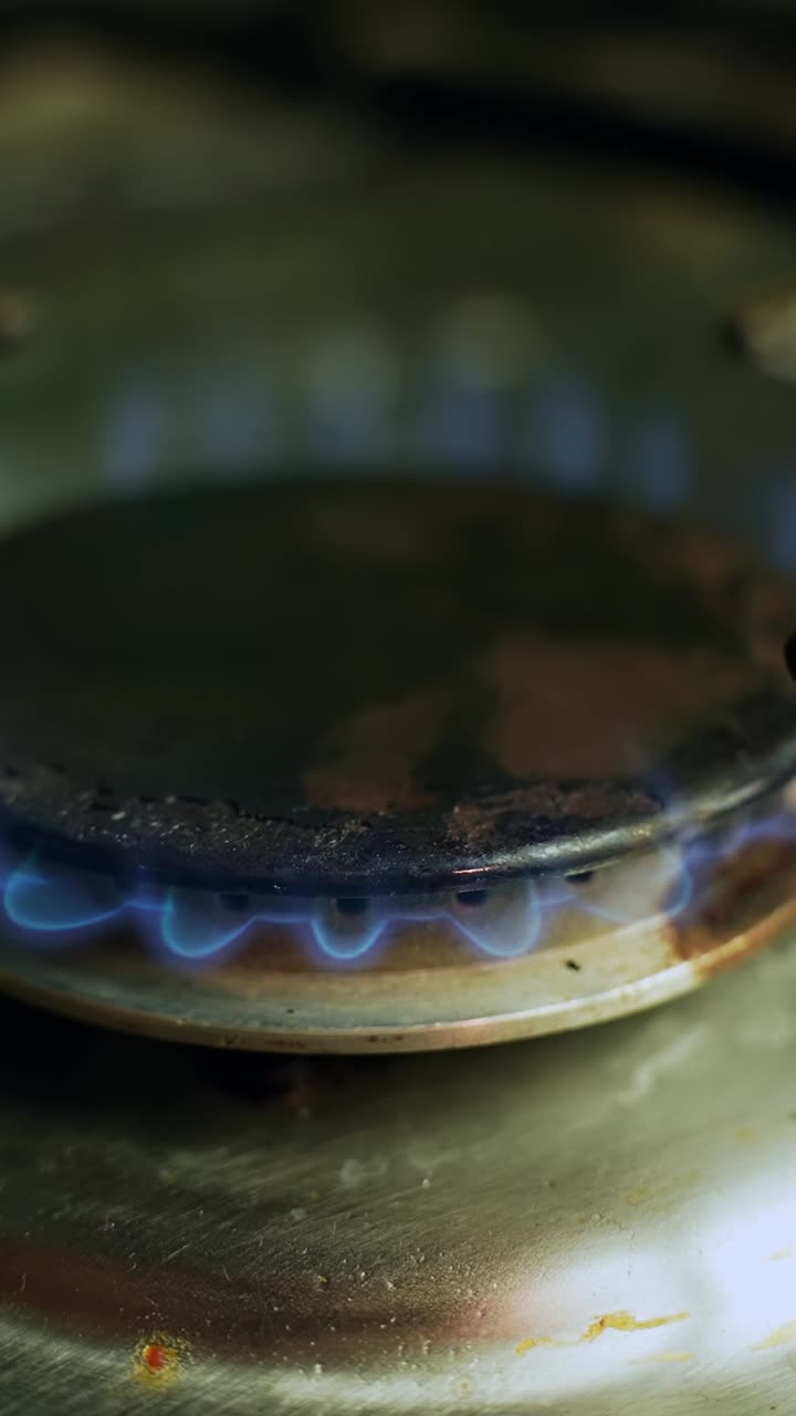 Close-up of a gas stove burner ignited with blue flame on a stainless steel cooktop, showing kitchen heat and energy. Vertical macro b-roll with copy space