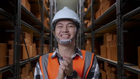 close up of asian male engineer with safety helmet standing in the warehouse with shelves full of delivery goods. smiling to the camera and clapping hands in the storage