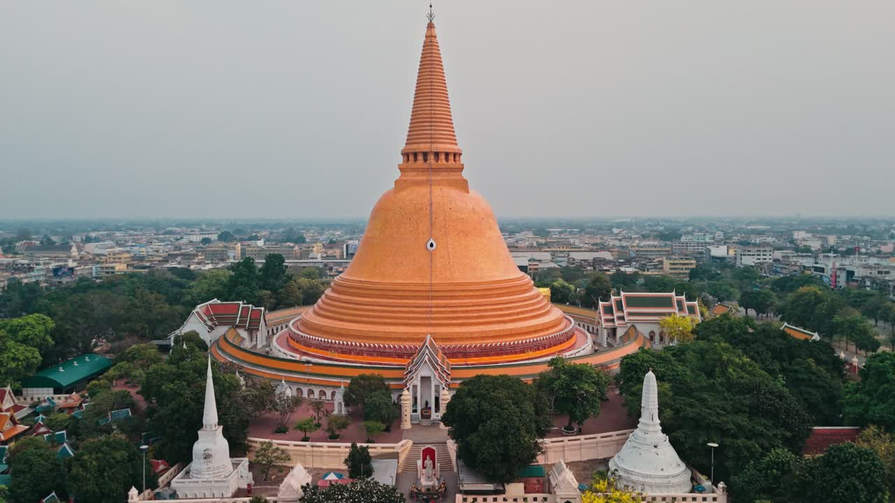 La prospettiva del drone orbita dolcemente verso destra mantenendo la messa a fuoco sulla gigantesca stupa arancione dorata mentre la luce nebbiosa del mattino inonda il lussureggiante cortile verde del tempio circondato da tetti di città estesi.