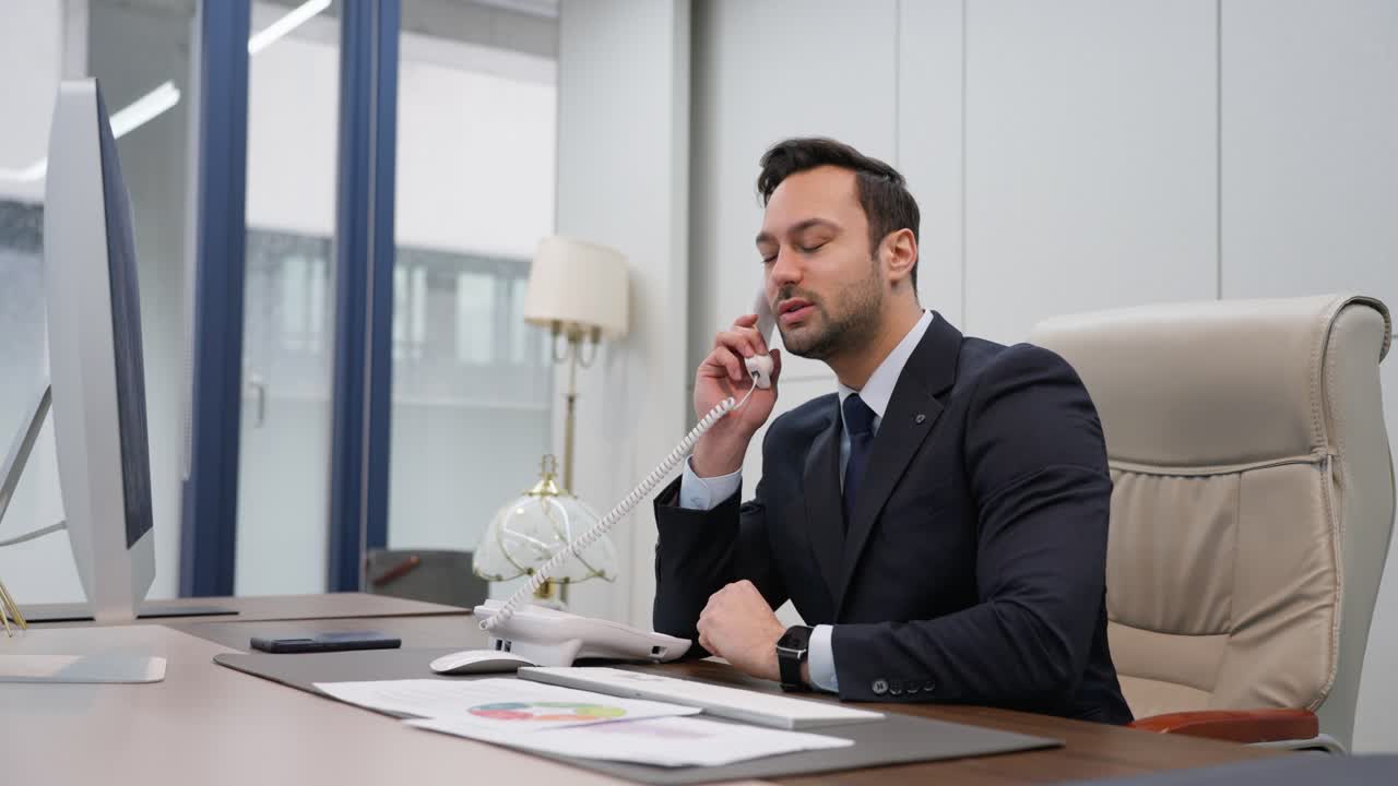 A confident professional in a suit is seated at his desk in a modern office, speaking on a landline phone while reviewing documents, exuding focus and determination