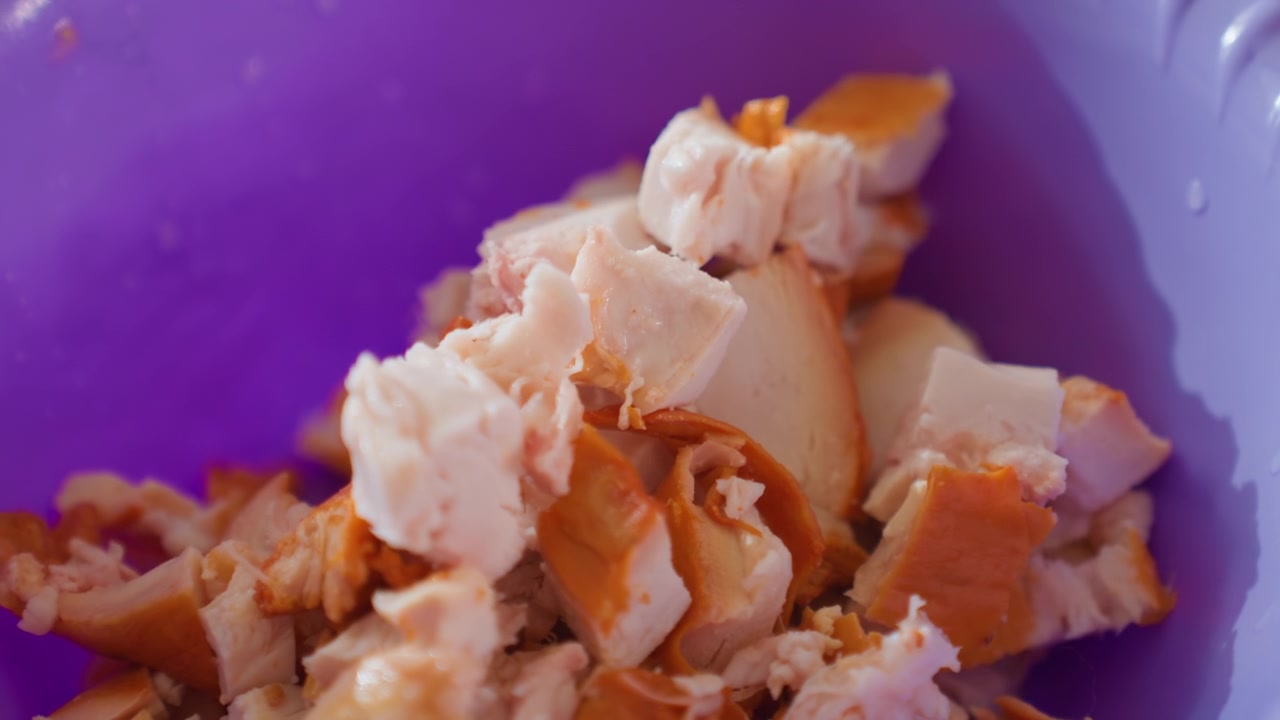 Primo piano di pollo cotto sfilacciato mescolato e sfilacciato con una forchetta di metallo all'interno di una ciotola di plastica viola brillante, con movimento visibile, consistenza e pezzi di carne sparsi durante la preparazione del cibo.