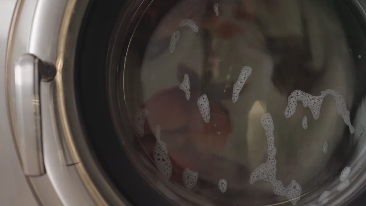 Close up on fabric swirling inside washing machine drum with soap suds forming on glass door, wet garments spinning during wash cycle, stainless steel appliance creating smooth motion in laundromat