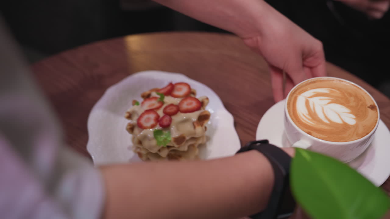 Close up of waitress wearing smart watch holding white ceramic cup of latte with elegant leaf-shaped milk foam art, serving hot beverage to customer in cozy indoor setting with waffles on table nearby